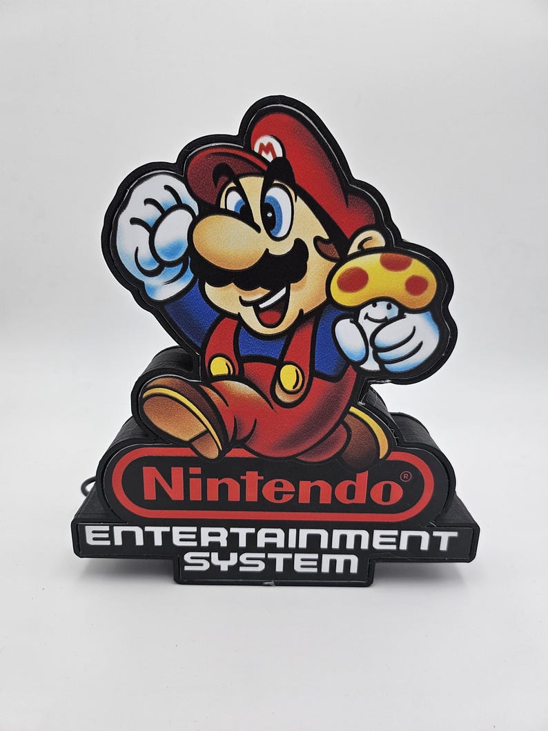 May include: A decorative light featuring Mario holding a yellow mushroom. The base is black with the red Nintendo logo and the white text ENTERTAINMENT SYSTEM. The character is depicted in a classic red and blue outfit with a brown hat and moustache.