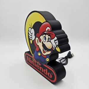 May include: A black, three-dimensional decorative sign featuring a graphic of Mario in a red hat and blue overalls. The sign includes a yellow crescent shape behind the character and a red oval base with the word Nintendo printed in black text.