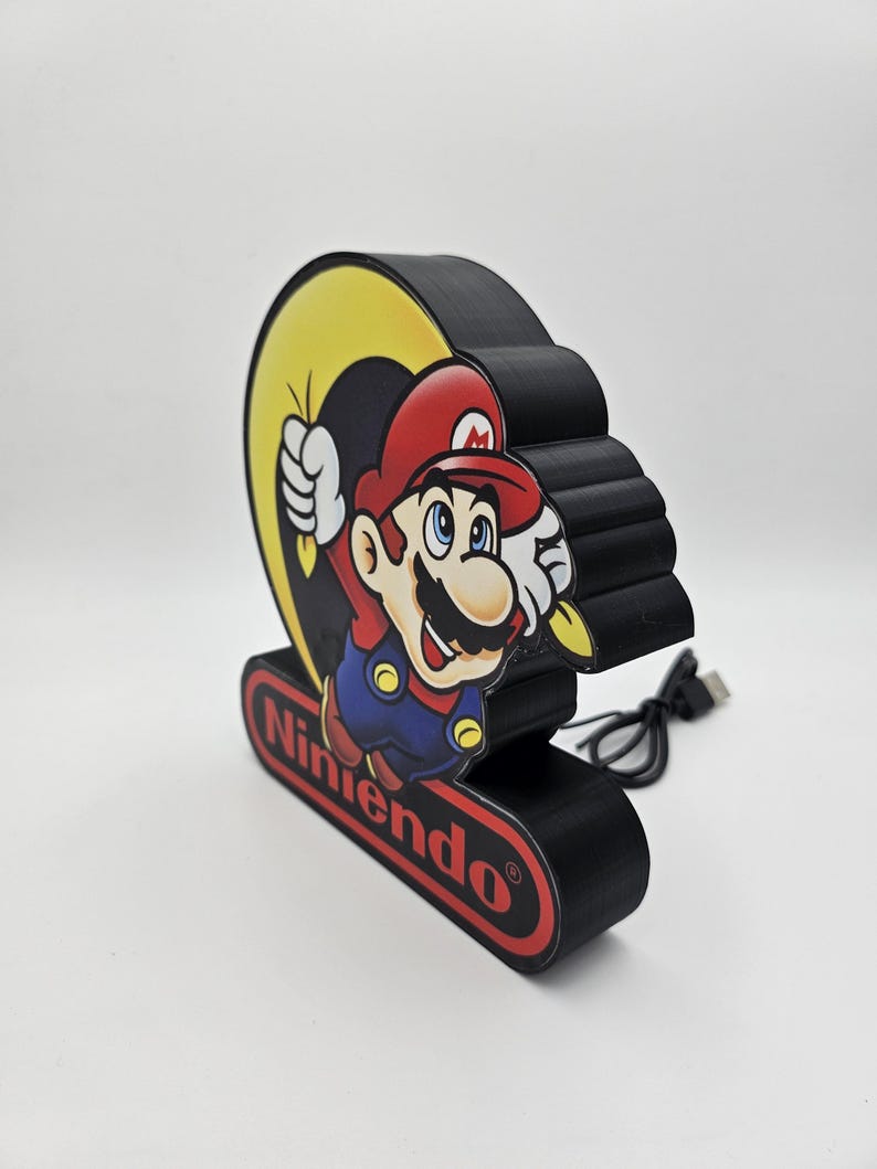 May include: A black, three-dimensional decorative sign featuring a graphic of Mario in a red hat and blue overalls. The sign includes a yellow crescent shape behind the character and a red oval base with the word Nintendo printed in black text.