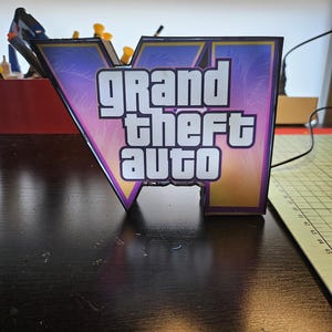 May include: A V-shaped sign featuring the Grand Theft Auto logo with a gradient background of purple, pink, and orange palm tree silhouettes. The text Grand Theft Auto is printed in bold white letters with a thick black outline on the front surface.