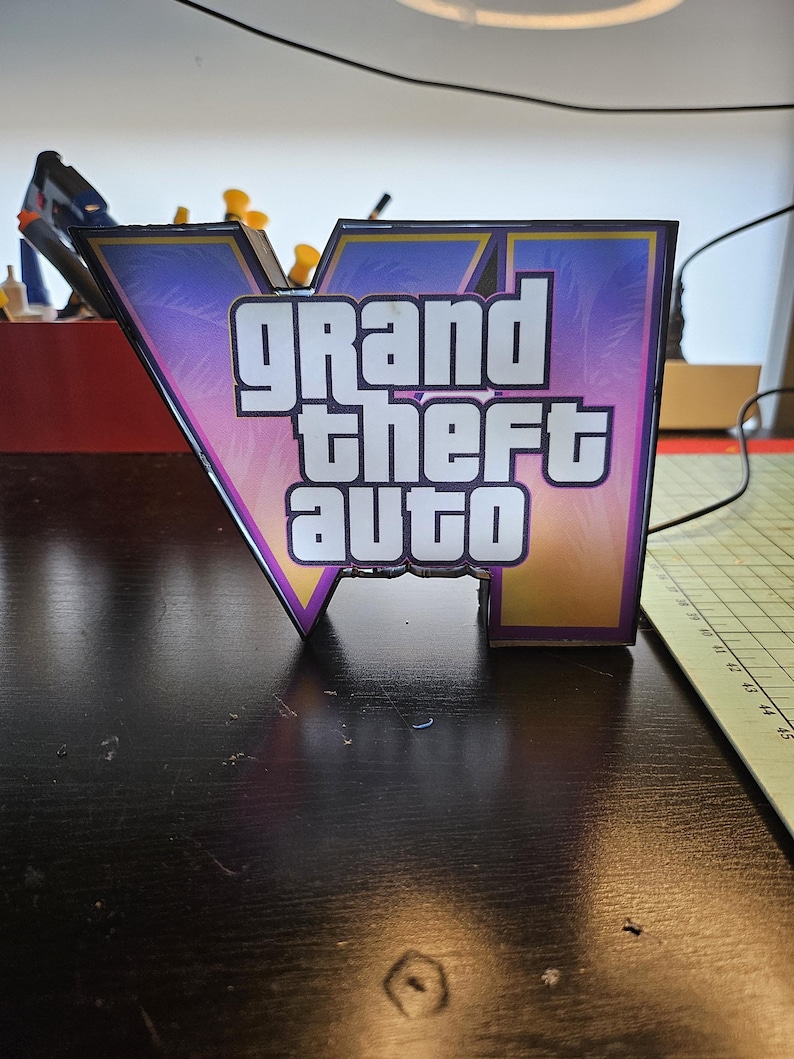 May include: A V-shaped sign featuring the Grand Theft Auto logo with a gradient background of purple, pink, and orange palm tree silhouettes. The text Grand Theft Auto is printed in bold white letters with a thick black outline on the front surface.