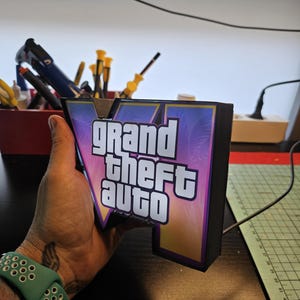May include: A three-dimensional light box sign featuring the Grand Theft Auto V logo. The sign has a black frame and a front panel with a purple and orange gradient background, palm tree silhouettes, and the white text Grand Theft Auto V.