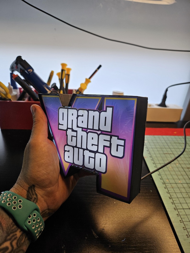 May include: A three-dimensional light box sign featuring the Grand Theft Auto V logo. The sign has a black frame and a front panel with a purple and orange gradient background, palm tree silhouettes, and the white text Grand Theft Auto V.