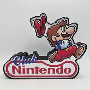 Puede incluir: Un soporte decorativo con un gr&aacute;fico de Mario corriendo y sosteniendo una gorra roja. La base tiene forma de &oacute;valo blanco con borde rojo y el texto Club Nintendo en letras azules y rojas. El objeto tiene un borde negro en capas.