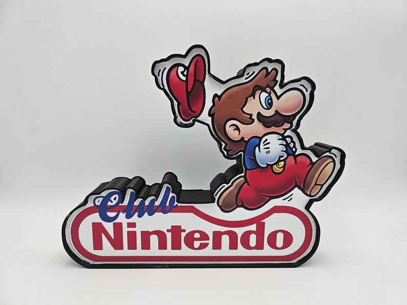 Puede incluir: Un soporte decorativo con un gr&aacute;fico de Mario corriendo y sosteniendo una gorra roja. La base tiene forma de &oacute;valo blanco con borde rojo y el texto Club Nintendo en letras azules y rojas. El objeto tiene un borde negro en capas.