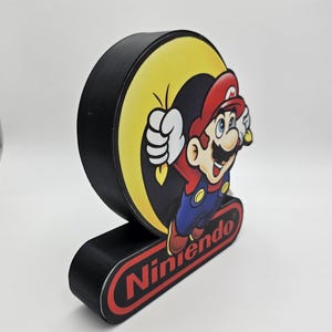 May include: A black, circular, three-dimensional display sign featuring a graphic of Mario jumping in front of a yellow circle. Below the circular graphic, a black rectangular base displays the red Nintendo logo with white text.