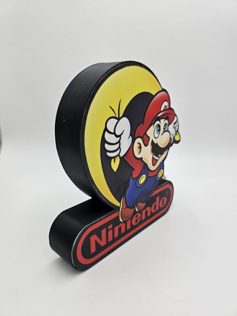 May include: A black, circular, three-dimensional display sign featuring a graphic of Mario jumping in front of a yellow circle. Below the circular graphic, a black rectangular base displays the red Nintendo logo with white text.