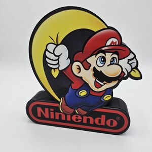 May include: A decorative light featuring the character Mario from Nintendo. Mario is depicted in his red cap, blue overalls, and white gloves, jumping in front of a yellow crescent moon. The base is a black rectangular block with the Nintendo logo.