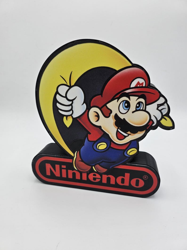 May include: A decorative light featuring the character Mario from Nintendo. Mario is depicted in his red cap, blue overalls, and white gloves, jumping in front of a yellow crescent moon. The base is a black rectangular block with the Nintendo logo.