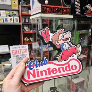Puede incluir: Un letrero de Club Nintendo que presenta un personaje de dibujos animados con cabello casta&ntilde;o, bigote, camisa azul y overol rojo, corriendo mientras sostiene una gorra roja. El letrero tiene un fondo blanco con bordes rojos y texto azul.