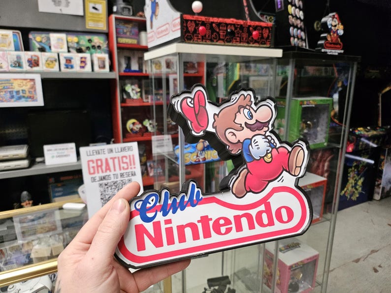 Puede incluir: Un letrero de Club Nintendo que presenta un personaje de dibujos animados con cabello casta&ntilde;o, bigote, camisa azul y overol rojo, corriendo mientras sostiene una gorra roja. El letrero tiene un fondo blanco con bordes rojos y texto azul.