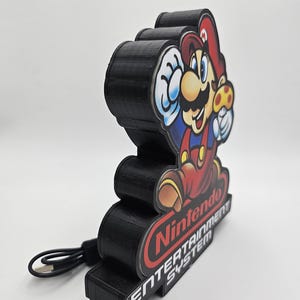 May include: A black 3D-printed display stand featuring a colourful graphic of Mario holding a mushroom. The base is labelled with the red Nintendo logo and the white text ENTERTAINMENT SYSTEM. A black USB power cable is attached to the side of the unit.