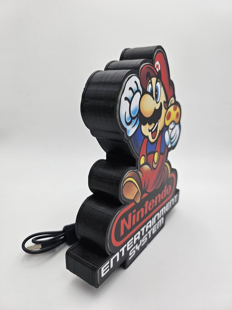 May include: A black 3D-printed display stand featuring a colourful graphic of Mario holding a mushroom. The base is labelled with the red Nintendo logo and the white text ENTERTAINMENT SYSTEM. A black USB power cable is attached to the side of the unit.