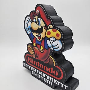 May include: A three-dimensional display piece featuring a colourful graphic of Mario holding a yellow mushroom. Below the character, the red Nintendo logo is visible, followed by the white text "ENTERTAINMENT SYSTEM" on a black base.