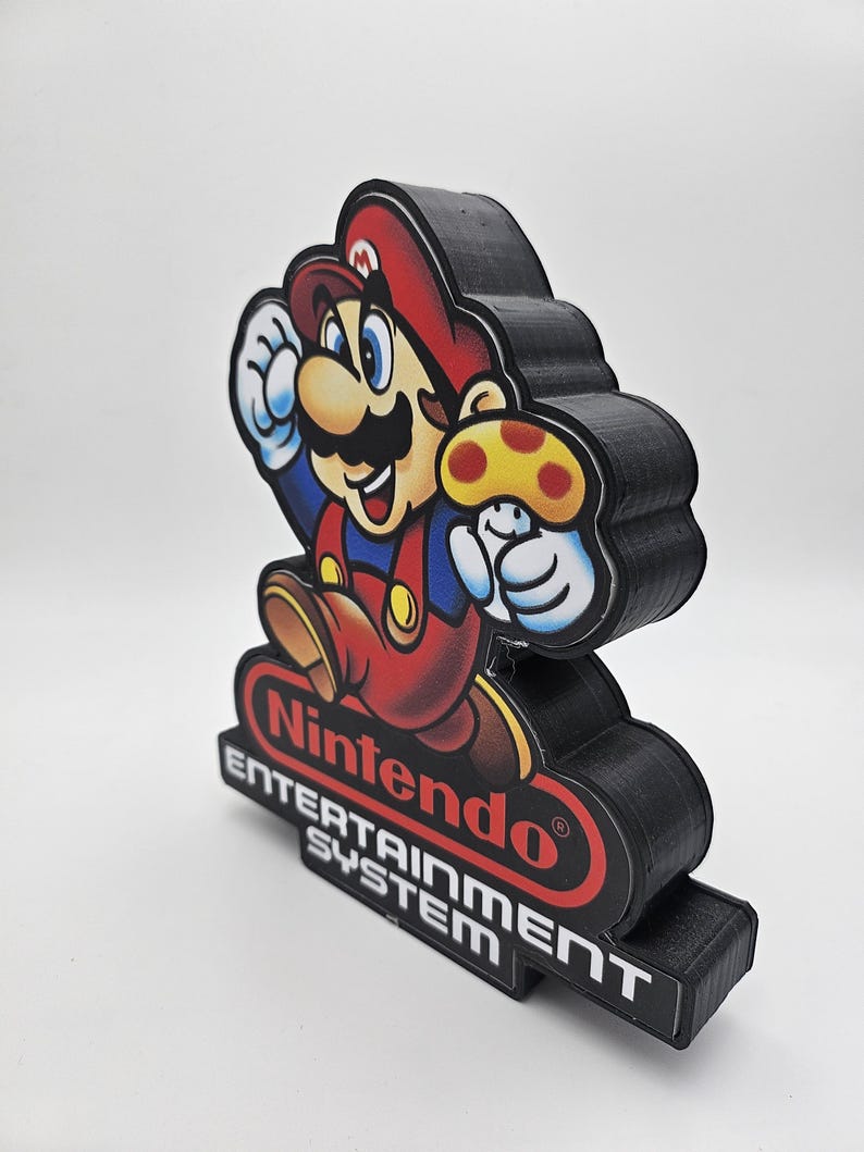 May include: A three-dimensional display piece featuring a colourful graphic of Mario holding a yellow mushroom. Below the character, the red Nintendo logo is visible, followed by the white text "ENTERTAINMENT SYSTEM" on a black base.