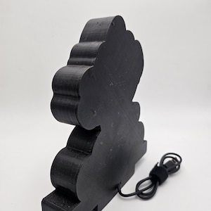 May include: A black, three-dimensional, cloud-shaped object with a layered, ridged texture. The object features a flat base and a black power cord extending from the lower back section, ending in a standard USB connector for electrical power.