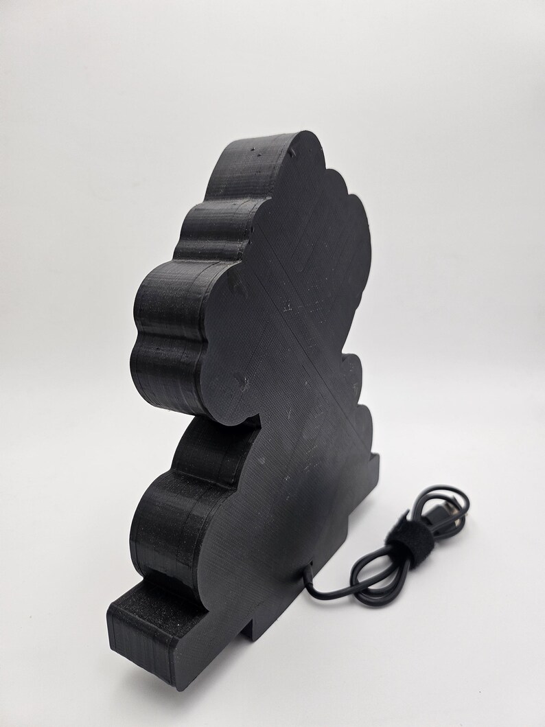 May include: A black, three-dimensional, cloud-shaped object with a layered, ridged texture. The object features a flat base and a black power cord extending from the lower back section, ending in a standard USB connector for electrical power.