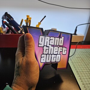 May include: A three-dimensional sign featuring the Grand Theft Auto V logo with a black frame and a gradient background of purple, pink, and orange. The white text Grand Theft Auto is centered on the front of the V-shaped structure.