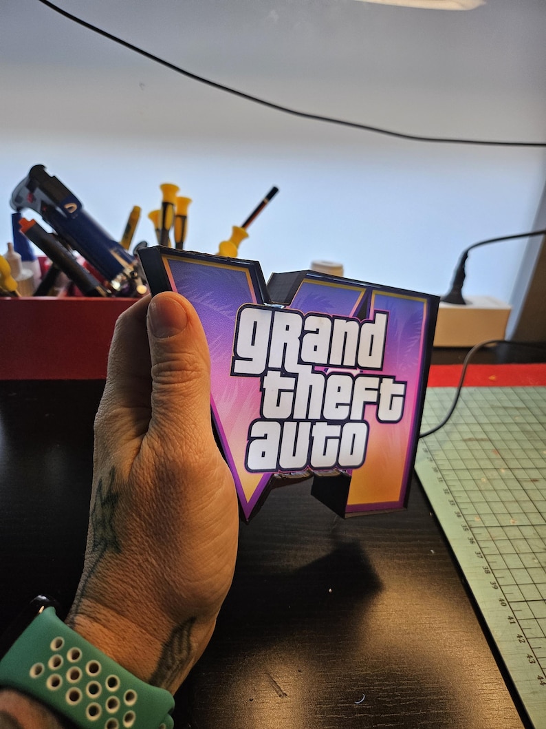 May include: A three-dimensional sign featuring the Grand Theft Auto V logo with a black frame and a gradient background of purple, pink, and orange. The white text Grand Theft Auto is centered on the front of the V-shaped structure.