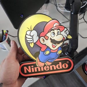May include: A decorative sign featuring a colorful illustration of Mario from Nintendo, depicted with a red hat, blue overalls, and a yellow sack. The sign includes the red and black Nintendo logo at the bottom, with a glossy finish and a die-cut shape.