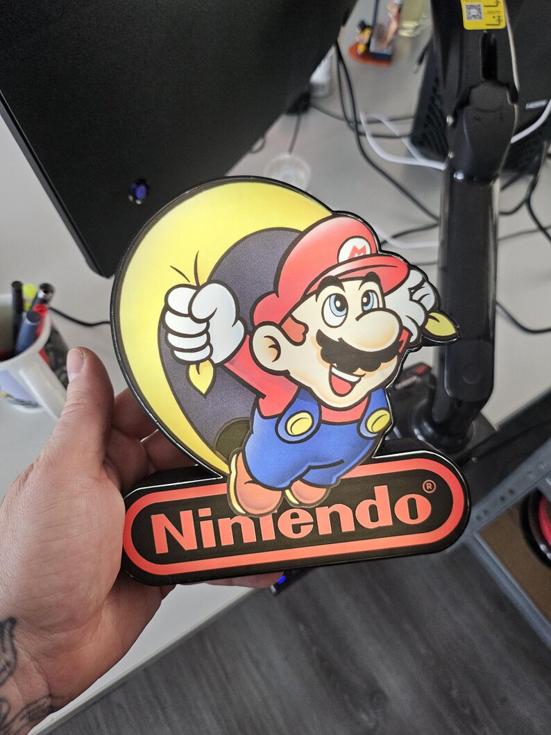 May include: A decorative sign featuring a colorful illustration of Mario from Nintendo, depicted with a red hat, blue overalls, and a yellow sack. The sign includes the red and black Nintendo logo at the bottom, with a glossy finish and a die-cut shape.