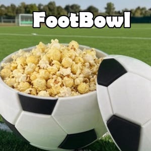 May include: A white and black soccer ball-shaped bowl filled with yellow popcorn, featuring a matching removable lid resting beside it. The text "FootBowl" is printed in white letters with a black outline above the bowl.