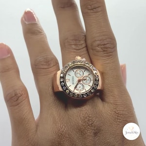 May include: A rose gold-toned ring watch featuring a round face with a white dial, the brand name BOZHI, and three small sub-dials. The bezel is encrusted with clear, sparkling crystals, and the band is a solid, polished rose gold-colored metal.
