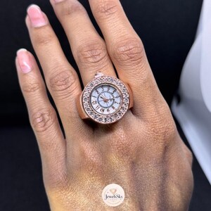 May include: A rose gold-tone ring watch featuring a round face with a white dial, black Arabic numerals, and a bezel encrusted with clear crystals. The watch crown is positioned at the top of the circular case, which is set on a wide metal band.