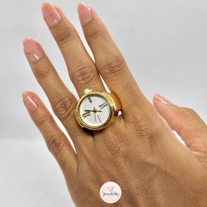 May include: A gold-toned ring featuring a functional watch face design. The circular dial is white with black Roman numeral markers at the twelve, three, six, and nine o'clock positions, and a small green and red stripe detail at the top.