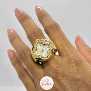 Puede incluir: Un anillo reloj de tono dorado con una esfera en forma de trébol, fondo blanco, manecillas doradas y pequeñas piedras transparentes engastadas en el bisel. La palabra QUARTZ está impresa en la esfera. El aro es ancho y de metal dorado pulido.