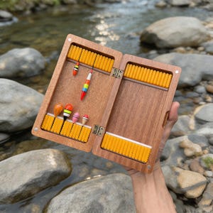 May include: A rectangular wooden fishing tackle box with a hinged design, featuring interior yellow foam inserts for organising gear. Five small, colourful fishing floats in orange, white, yellow, and pink are attached to the foam padding.