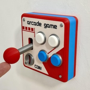 Op de afbeelding: Een vierkante arcade game lichtschakelaar voor aan de muur met een rood-witte voorplaat met de tekst arcade game en coin. Het bevat een zilveren joystick met een rode knop, twee witte knoppen en twee blauwe knoppen, omlijst door een blauwe rand.