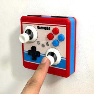 Op de afbeelding: Een vierkante lichtschakelaarafdekking ontworpen als een retro gamecontroller. Het heeft een witte voorzijde met rode en blauwe strepen, het woord Gamepad, een zwart richtingspad, twee witte joysticks en rode X, Y en blauwe A, B knoppen.