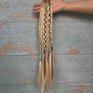 May include: A set of blonde hair extensions featuring multiple braided strands. The braids are accented with dark brown cord wrapping and small turquoise-colored beads. The hair strands are straight and hang vertically in a long, layered style.