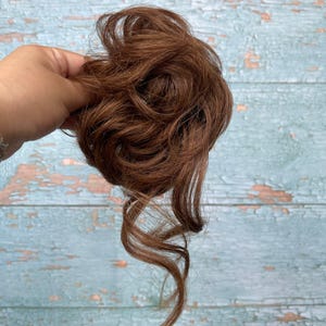 May include: A medium brown hair scrunchie extension featuring wavy, layered synthetic hair strands arranged in a voluminous, messy bun style. The hairpiece has a circular base with loose, curled tendrils extending downward from the main cluster.