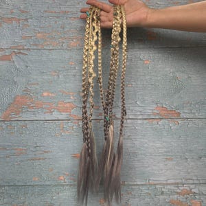 May include: A set of hair extensions featuring multiple blonde and light brown braided strands. The braids incorporate various textures, including twisted sections and loose ends, accented with small turquoise beads and metallic star-shaped charms.