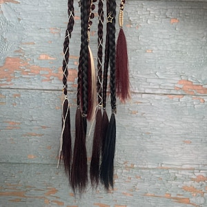 Può includere: Un set di extension per capelli con diverse trecce marrone scuro e nere. Le trecce sono decorate con sottili fili bianchi e dorati, un piccolo ciondolo a forma di stella dorata e punte di capelli sciolte e lisce in bordeaux e nero.