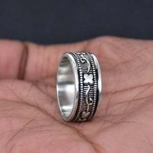 Könnte beinhalten: Ein silberfarbener Metallring mit einem breiten Band und oxidiertem Finish. Die Oberfläche zeigt ein sich wiederholendes Muster aus kleinen vierblättrigen Blumenformen und vertikalen Linienstrukturen, eingefasst von feinen Perlenrändern.