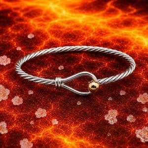 Könnte beinhalten: Ein Armreif aus gedrehtem, silberfarbenem Kabel mit einem Verschluss im Haken-Stil. Der Verschluss besteht aus einer glatten, polierten goldfarbenen Metallkugel, die an einer geschwungenen silberfarbenen Schlaufe befestigt ist.