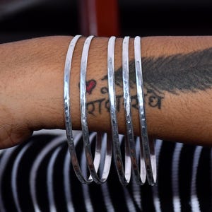 Könnte beinhalten: Ein Set aus fünf dünnen, silberfarbenen Armreifen mit einer gehämmerten, strukturierten Oberfläche. Die Armreifen sind kreisförmig und haben eine reflektierende, metallische Oberfläche, die das Licht einfängt und parallele Linien bildet.