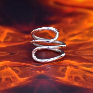 Könnte beinhalten: Ein silberfarbener Ring mit einem einzigartigen, offenen Design. Der Ring hat zwei längliche, gebogene Schlaufen, die sich um den Finger wickeln. Das Metall hat eine polierte Oberfläche, die das Licht reflektiert. Der Ring ist vor einem feurigen, orange-schwarzen Hintergrund platziert.