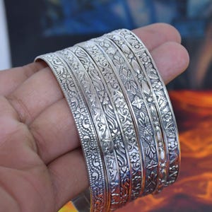 Könnte beinhalten: Ein Set aus sechs silberfarbenen Metallarmreifen mit detaillierten floralen und geometrischen Prägemustern. Jeder Armreif weist ein individuelles Reliefdesign auf seiner Oberfläche auf und zeigt ein poliertes Metallfinish mit abgedunkelten Vertiefungen.