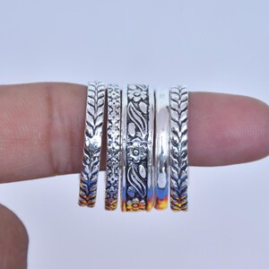 Könnte beinhalten: Ein Set aus fünf silberfarbenen Ringen mit verschiedenen Texturen und Mustern, darunter zwei Ringe mit einem sich wiederholenden Blattdesign, einer mit einem kleinen geometrischen Muster, einer mit Blumenmotiv und ein glattes, poliertes Band.