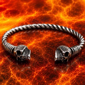 Könnte beinhalten: Ein Armreif aus silberfarbenem Metall mit einem gedrehten, seilartigen Band, das an beiden Enden mit einem detaillierten Totenkopf-Ornament abschließt. Der Armreif hat ein offenes Design, sodass er einfach über das Handgelenk gestreift werden kann.