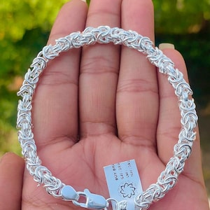 May include: A silver Byzantine chain bracelet featuring a polished finish and a lobster claw clasp. A small white rectangular tag is attached to the clasp, displaying the text 'BOY BR', 'GW: 24.0', 'LVV:', and 'NW: 24.0' along with a small floral icon.
