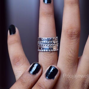Könnte beinhalten: Ein Stapel aus fünf Silberringen an einem Finger. Das Set besteht aus Ringen mit unterschiedlichen Texturen, darunter ein Blumenmuster, ein Perlendesign, ein gedrehtes Seildetail, ein glatt poliertes Band und ein gerillter Rand für einen Lagen-Look.