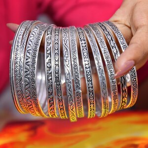 Könnte beinhalten: Eine Sammlung von silberfarbenen Metallarmreifen mit verschiedenen komplizierten Prägemustern, darunter florale Motive, geometrische Formen, Sterne und Herzdesigns, die in einer gestapelten Formation mit poliertem und oxidiertem Finish angeordnet sind.
