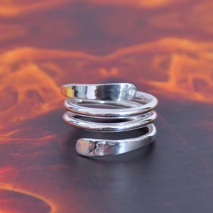Könnte beinhalten: Ein silberfarbener Metallring mit einem mehrschichtigen Spiraldesign. Das Band besteht aus drei durchgehenden Windungen, die sich um den Finger legen, mit abgeflachten, leicht texturierten Enden an der Ober- und Unterseite der Spirale.