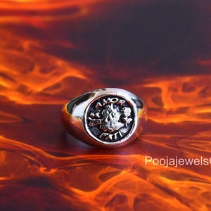 Peut inclure: Une bague chevalière argentée avec un plateau circulaire orné d'un motif en relief et de l'inscription AMOR FATI. La bague présente une finition métallique polie et un anneau lisse et arrondi. Le motif central est entouré de détails gravés.