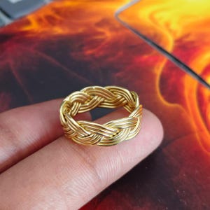 Könnte beinhalten: Ein goldfarbener Ring mit einem geflochtenen Drahtdesign. Das Band besteht aus mehreren dünnen, metallischen Strängen, die in einem durchgehenden, sich wiederholenden Muster miteinander verflochten sind und eine texturierte, seilartige Optik erzeugen.